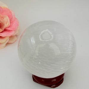 May include: A spherical white selenite crystal ball with a fibrous, translucent texture resting on a small, circular dark brown wooden stand. A large, soft pink artificial flower is positioned to the left of the crystal sphere.