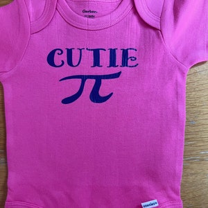 Puede incluir: Un body de beb&eacute; rosa con la palabra "CUTIE" y el s&iacute;mbolo pi en azul marino. El body de manga corta tiene un cuello redondo. La marca "Gerber" est&aacute; impresa en el cuello.