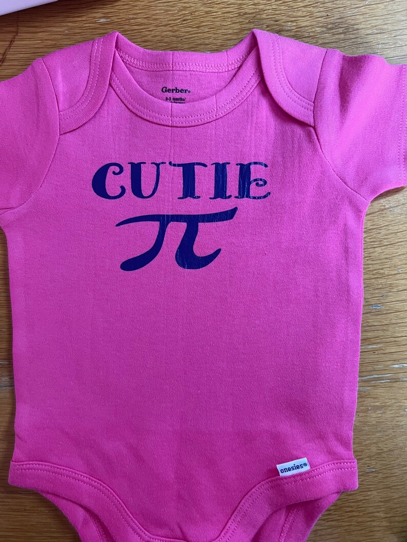 Puede incluir: Un body de beb&eacute; rosa con la palabra "CUTIE" y el s&iacute;mbolo pi en azul marino. El body de manga corta tiene un cuello redondo. La marca "Gerber" est&aacute; impresa en el cuello.