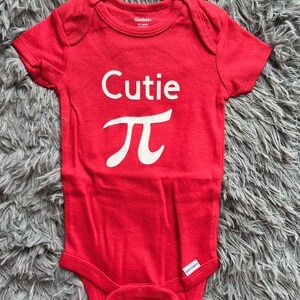 Puede incluir: Un body rojo para beb&eacute; con la palabra "Cutie" y el s&iacute;mbolo matem&aacute;tico pi en blanco. El body de manga corta tiene cierres de presi&oacute;n en la parte inferior. Las marcas "Gerber" y "onesies" son visibles. Un atuendo lindo.