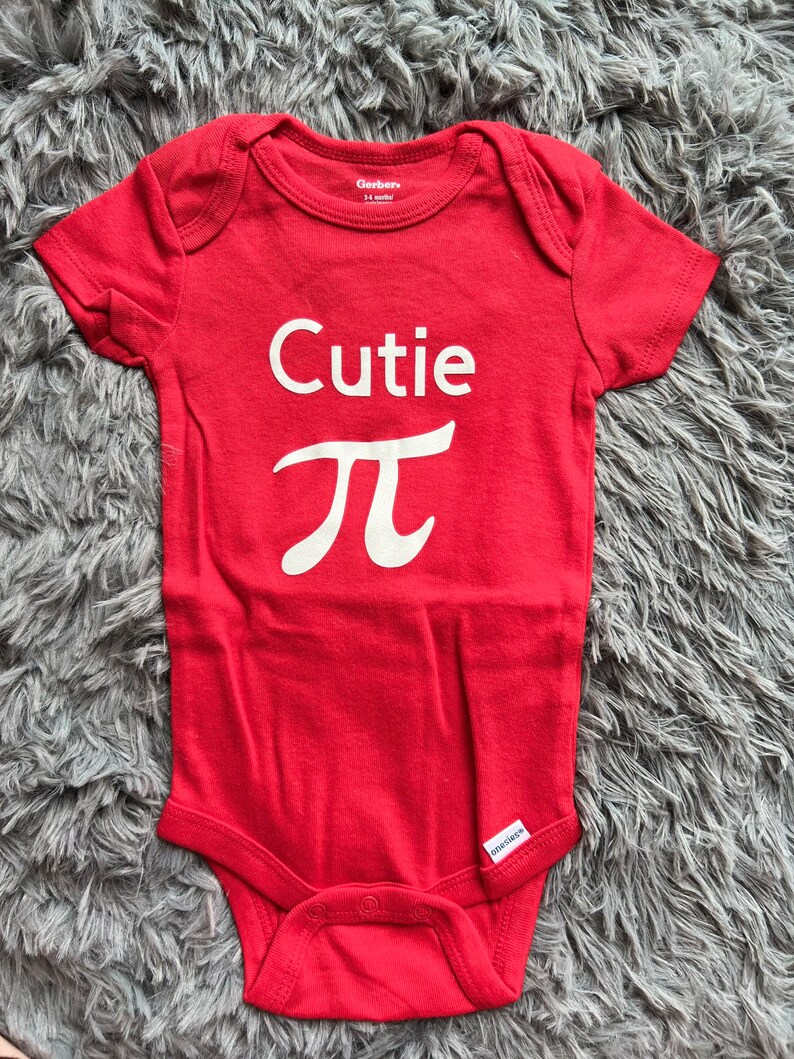 Puede incluir: Un body rojo para beb&eacute; con la palabra "Cutie" y el s&iacute;mbolo matem&aacute;tico pi en blanco. El body de manga corta tiene cierres de presi&oacute;n en la parte inferior. Las marcas "Gerber" y "onesies" son visibles. Un atuendo lindo.