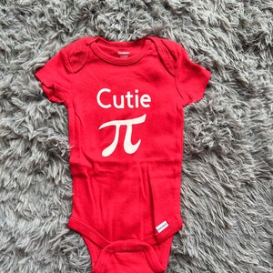 Puede incluir: Un body rojo para beb&eacute; con la palabra "Cutie" y el s&iacute;mbolo pi en blanco. El body est&aacute; hecho de tela suave y tiene mangas cortas. La etiqueta de la marca Gerber es visible.