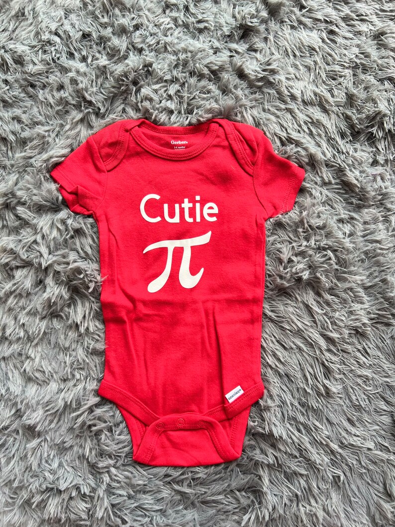 Puede incluir: Un body rojo para beb&eacute; con la palabra "Cutie" y el s&iacute;mbolo pi en blanco. El body est&aacute; hecho de tela suave y tiene mangas cortas. La etiqueta de la marca Gerber es visible.