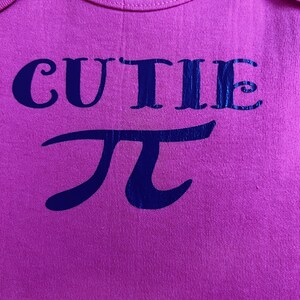 Puede incluir: Body rosa para beb&eacute; con la palabra "CUTIE" y el s&iacute;mbolo matem&aacute;tico pi en azul oscuro. La prenda es de tejido suave y tiene cuello redondo. La etiqueta indica la marca Gerber y la talla.