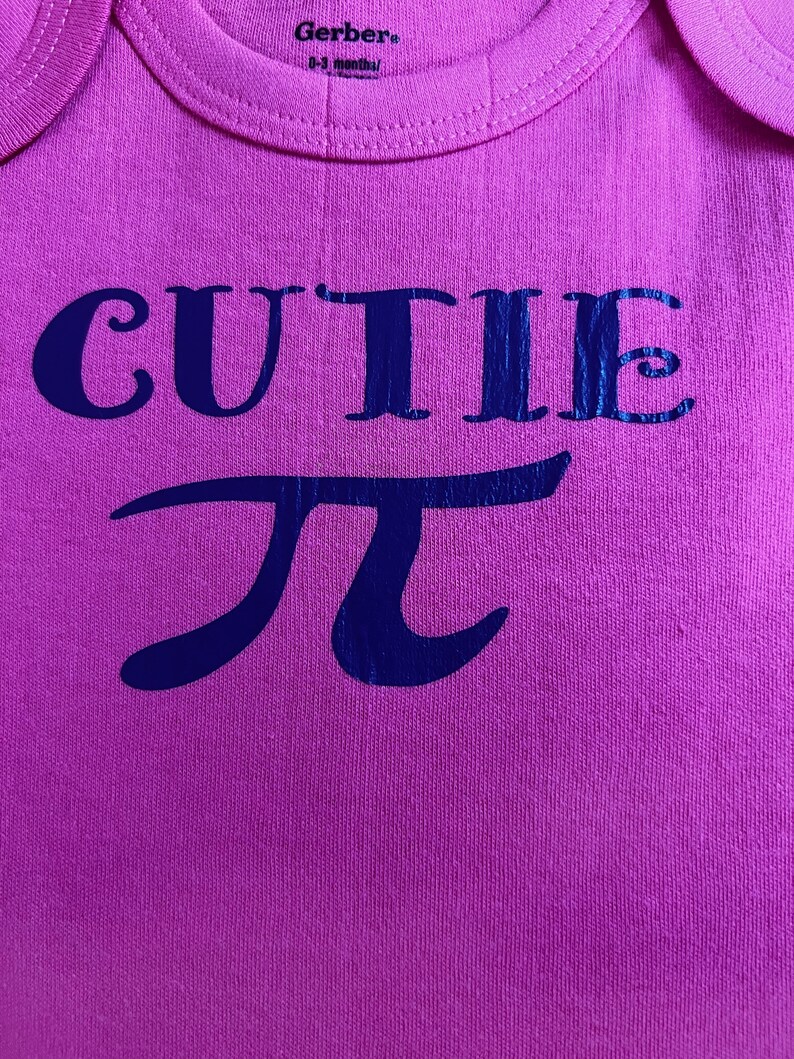Puede incluir: Body rosa para beb&eacute; con la palabra "CUTIE" y el s&iacute;mbolo matem&aacute;tico pi en azul oscuro. La prenda es de tejido suave y tiene cuello redondo. La etiqueta indica la marca Gerber y la talla.