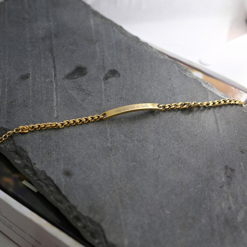 May include: A gold-coloured bracelet with a curb chain design. The bracelet has a rectangular bar engraved with the text "You had me at hello". The jewellery is displayed on a dark grey, textured surface.