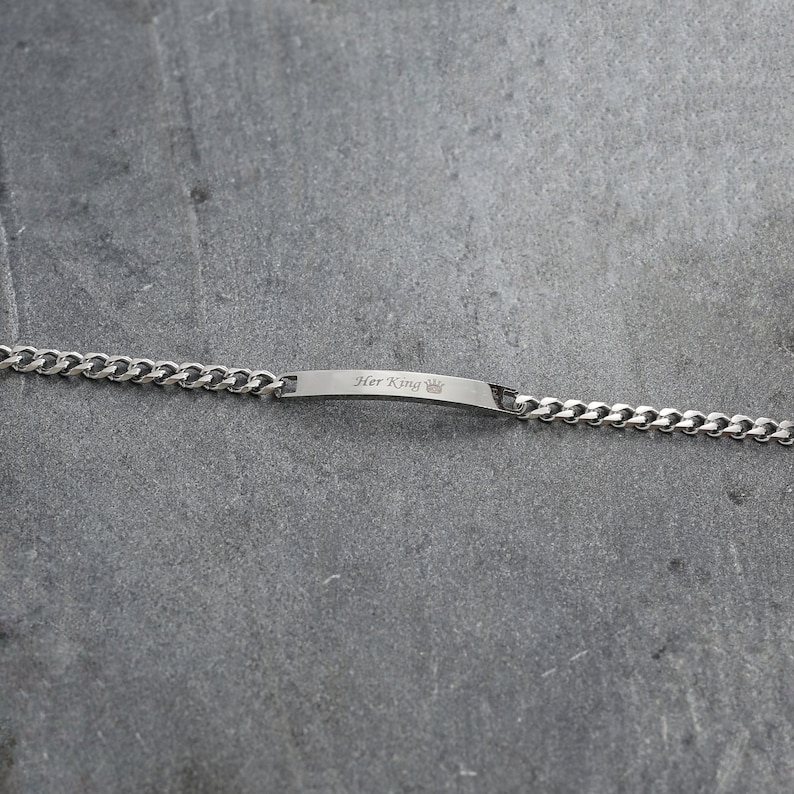 May include: A silver bracelet with a curb chain. The rectangular bar is engraved with the words "Her King" and a crown symbol. The bracelet is shown on a grey surface.