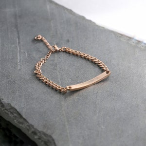 May include: Rose gold bracelet featuring a curb chain and a rectangular bar. The bar is engraved with the name "Logan" and two small hearts. The bracelet is shown on a grey surface.