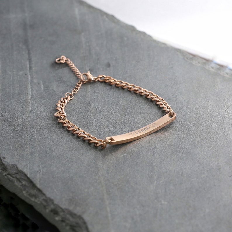 May include: Rose gold bracelet featuring a curb chain and a rectangular bar. The bar is engraved with the name "Logan" and two small hearts. The bracelet is shown on a grey surface.