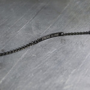 May include: A black bracelet with a chain link design. The bracelet features a rectangular bar with the words "STRONG ALONE" engraved on it. The bracelet is displayed on a grey surface.