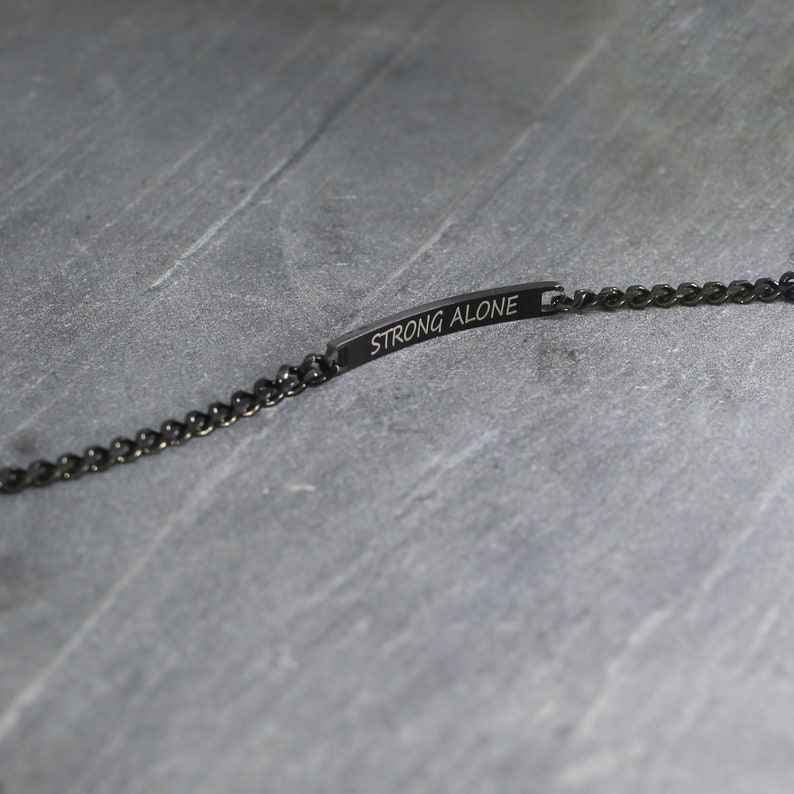 May include: A black bracelet with a chain link design. The bracelet features a rectangular bar with the words "STRONG ALONE" engraved on it. The bracelet is displayed on a grey surface.