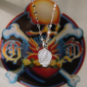 May include: A silver chain necklace with a silver skull pendant featuring a lightning bolt design. The necklace is hanging in front of a poster with a skull and crossbones graphic.