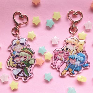 May include: Two gold keychains with clear acrylic charms featuring anime characters. The first charm features three girls with pink, green, and blue hair. The second charm features three girls with yellow, pink, and blue hair.