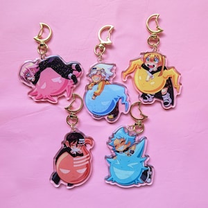 May include: Five acrylic keychains featuring anime-style characters paired with colourful creatures. Each charm includes a gold-toned crescent moon clasp. The designs include characters with pink, white, yellow, black, and blue colour schemes.