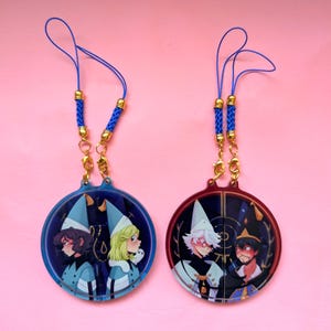 May include: Two circular acrylic charms featuring anime-style character illustrations. The left charm has blue tones with two figures in light blue hats. The right charm has red tones with two figures in dark hats. Both include blue braided cords.