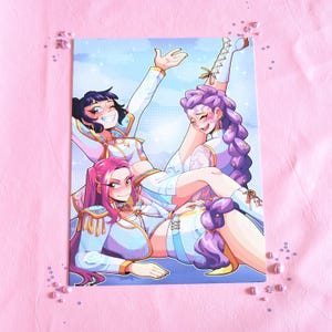 May include: A print featuring three stylized figures in white and light blue outfits with gold accents. One figure has black hair, another has pink hair, and the third has purple hair. The background is light blue with stars.