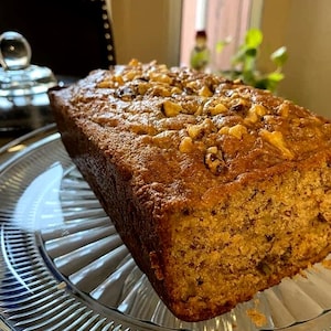 May include: A loaf of banana bread with a golden-brown crust, topped with chopped walnuts, sits on a clear, ridged glass plate. The bread has a moist appearance, suggesting a freshly baked treat. The background is slightly blurred, with a glimpse of a window and a decorative object.