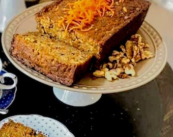 May include: A sliced loaf of carrot cake on a decorative plate, garnished with shredded carrots and walnuts. The cake is on a white pedestal cake stand. A slice of cake is on a small plate with a blue and white pattern.