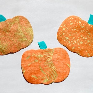 May include: Three orange paper pumpkins with gold glitter accents and teal stems. The pumpkins are arranged in a triangular pattern on a white background.