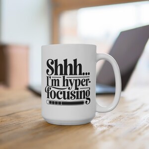 Op de afbeelding: Witte keramische mok met een groot handvat. De mok heeft de tekst "Shhh... I'm hyper-focusing" in het zwart, met een laadbalk-afbeelding. De mok staat op een houten oppervlak, met een laptop op de achtergrond.
