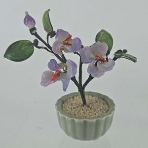 May include: A decorative bonsai-style sculpture featuring light purple glass flowers with bright red stamens and translucent green glass leaves. The branches are wrapped in dark thread, and the plant is set in a small, fluted, sage green ceramic pot filled with light-colored gravel.
