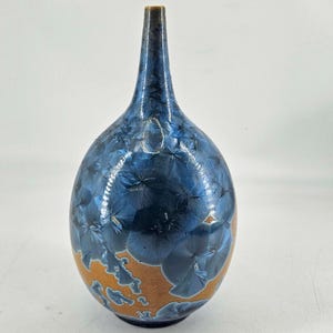 May include: A decorative ceramic vase with a dark blue crystalline glaze over a tan base. The vase has a long, slender neck and a rounded body. The glaze has a unique, textured appearance.