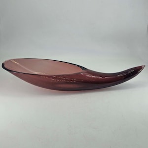 May include: A long, curved, decorative glass bowl in a deep plum color. The bowl has a smooth, glossy finish and a unique, elongated shape, perfect for displaying fruit or other decorative items. The bowl's design is both modern and elegant.
