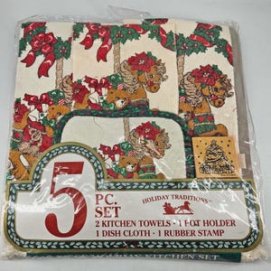 May include: A five-piece Holiday Traditions kitchen set in a clear plastic package. The set includes two kitchen towels, a pot holder, a dish cloth, and a rubber stamp. The towels and pot holder feature a carousel horse and teddy bear design with red and green accents.