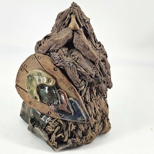 May include: A handcrafted ceramic figurine of a bearded warrior. The figure is primarily brown with a shield featuring metal accents in green, blue, and copper. The beard and hair are textured, adding detail to the sculpture.