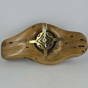 May include: A horizontal clock crafted from a piece of natural light brown wood with a smooth, polished finish. A circular brass faceplate with a black geometric pattern is centered, featuring white clock hands and a four-pointed brass frame.