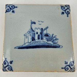 May include: Square ceramic tile with a blue and white illustration of a castle on a hill, a well, and birds. The corners feature decorative blue scrollwork. The tile has a slightly aged appearance.
