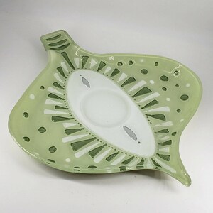May include: A light green ceramic dish shaped like a leaf with a central white oval section. The surface features dark green and white painted geometric patterns, including triangles, dots, and lines, surrounding a smooth, circular indentation.