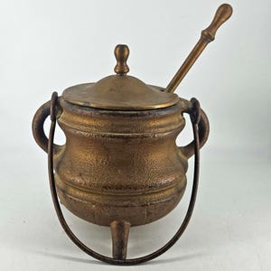 May include: A small, antique bronze-colored cauldron with a lid and a spoon. The cauldron has two handles and a curved handle. The cauldron is resting on three small legs. The spoon is resting on the lid.