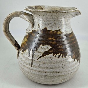 May include: A white ceramic pitcher with brown glaze accents. The pitcher has a rounded body and a handle. The glaze is applied in a drip pattern.