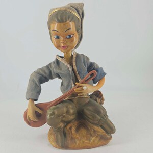 May include: A vintage-style figurine of a musician wearing a grey fabric jacket and matching headpiece with gold trim. The figure has painted facial features, blue eyes, and is seated while holding a brown stringed instrument with a long neck.