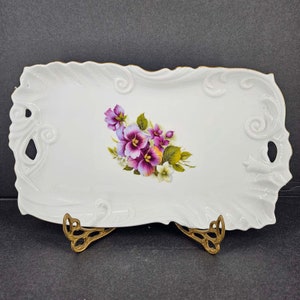 May include: A white porcelain serving tray with a floral design featuring purple and white pansies. The tray has a gold trim and ornate, scroll-like handles. The tray is supported by a gold-colored metal stand.
