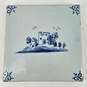 May include: Square ceramic tile with a hand-painted landscape scene in shades of blue. The design features a building, trees, and birds against a light blue background. Decorative scrollwork adorns the corners.