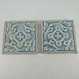 May include: Two square ceramic tiles featuring a raised relief pattern in shades of light blue and grey. The design consists of a central diamond shape with circular accents at each corner, surrounded by symmetrical scrolling leaf motifs.