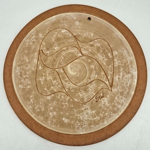 May include: Round ceramic wall hanging with a brown rim and a light brown center. The center features a swirling design with a carved floral pattern and the artist's signature, "Enid". A small hole is at the top for hanging.