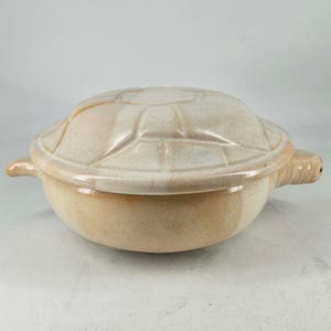 May include: A ceramic turtle-shaped casserole dish with a lid. The dish is a light beige color with a textured, segmented lid resembling a turtle shell. It has small handles and a spout-like feature.