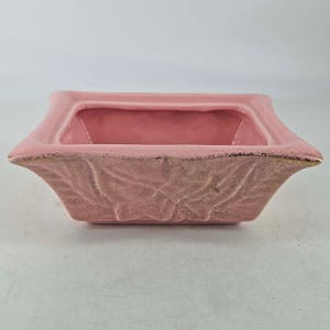 May include: A pink ceramic planter with a rectangular shape and a textured leaf design. The planter has a slightly flared rim with gold accents. The interior is a solid pink color, and the exterior has a matte finish.