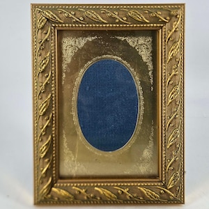 May include: A vintage gold-toned picture frame with an oval opening and a dark blue backing. The frame has a detailed leaf and vine design around the edges, adding a touch of elegance. The frame is rectangular and has a distressed finish.
