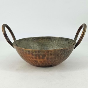 May include: A small, hammered copper bowl with two arched handles. The bowl has a rounded shape and a textured surface. The interior has a silver-gray color, while the exterior is a mix of copper and bronze tones.