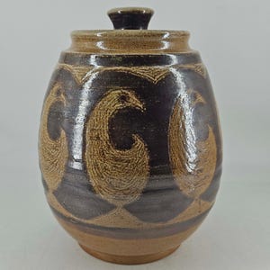 May include: A ceramic jar with a lid, featuring a dark brown glaze and a repeating pattern of light tan, textured bird silhouettes encircling the body. The vessel has a rounded shape, a short neck, and a matching lid with a small knob handle.