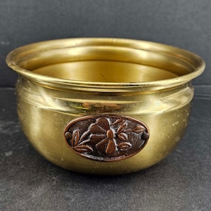 May include: A gold brass planter with a decorative copper and brass flower design on the front. The planter is round and has a slightly flared rim.