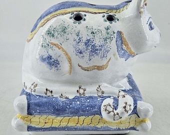 May include: A ceramic cat figurine with blue, yellow, and green painted details. The cat is sitting on a blue and yellow cushion.