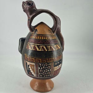 May include: A brown and black ceramic vessel with a handle and a figure on top. The vessel features geometric patterns in red, tan, and black. It has a small spout and sits on a round base. The handle curves over the top.