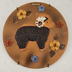 May include: A brown ceramic wall hanging with a black sheep with white spots on its head. The sheep is surrounded by colorful flowers and leaves.