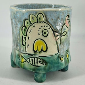 May include: Handmade ceramic planter with a whimsical fish design. The square-shaped planter features a light blue exterior with a green base and feet. The fish is cream-colored with yellow and green accents, and a pink flower.