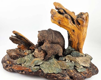 May include: A wooden sculpture of a mother bear and her cub nestled in a rocky, wooded setting. The sculpture is made of natural wood and features realistic details.